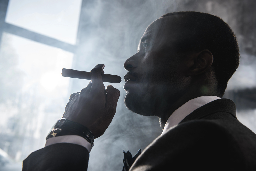 Man in suit smoking a cigar surrounded by smoke, dramatic low-angle silhouette with moody lighting